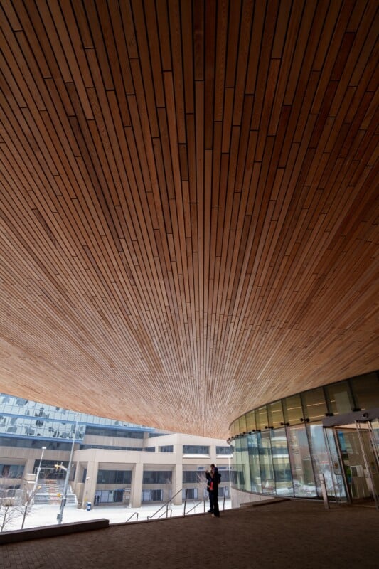 The image shows a wide, curved wooden ceiling with a pattern of parallel lines extending outward. A person stands underneath near a glass façade of a modern building. Snow is visible on the ground outside.