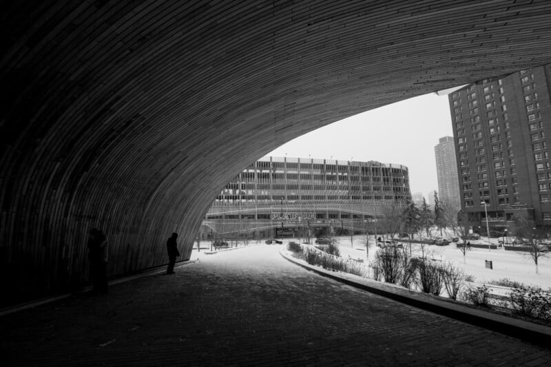 Black and white photo of a snowy urban scene viewed through a curved wooden structure. Two people stand in silhouette. In the background, there's a parking garage and tall buildings. Sparse snow-covered bushes line the path.
