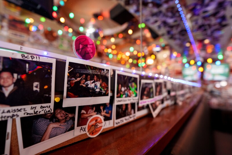 Close-up of a bar counter decorated with colorful string lights and a line of Polaroid photos displaying people in various poses. A ping pong ball with a signature is balanced atop the photos. The background is softly blurred.