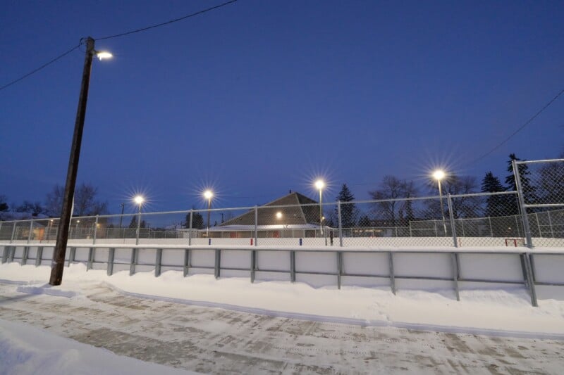 A snow-covered outdoor hockey rink is illuminated by tall floodlights against a darkening blue sky. A wooden building is visible in the background, surrounded by leafless trees and a chain-link fence.