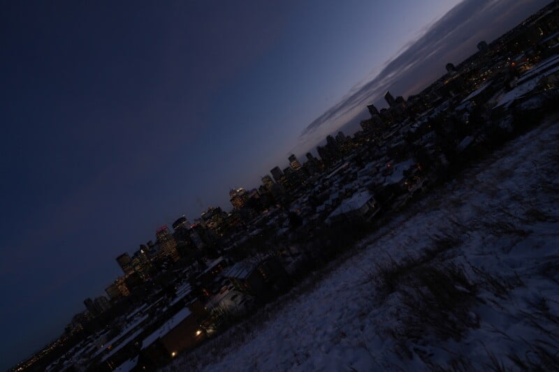 A city skyline at dusk viewed from a snowy hill. Buildings are illuminated against the darkening sky, with a mix of bright and faint lights. The foreground shows snow and sparse vegetation.