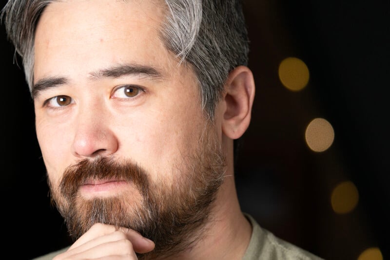 A man with salt-and-pepper hair and a beard poses with his hand touching his chin. He has a thoughtful expression. The background is dark with blurred circular lights.