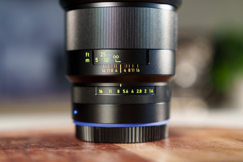 Close-up of a camera lens with focus and aperture scales visible. The lens shows measurements in feet and meters, and aperture values ranging from 2 to 16. The background is softly blurred.