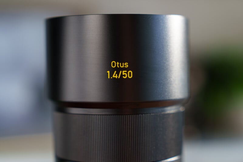 Close-up of a camera lens with "Otus 1.4/50" written in yellow on its barrel. The lens has a sleek, metallic finish and a ribbed grip section below the text. The background is softly blurred.