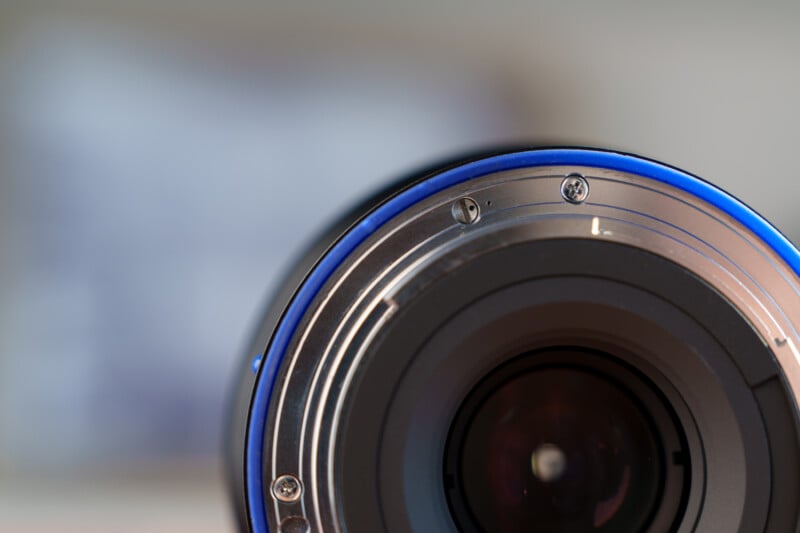 Close-up of a camera lens with a metallic and blue ring around its circumference. The background is blurred, highlighting the lens's intricate design and reflective surface.