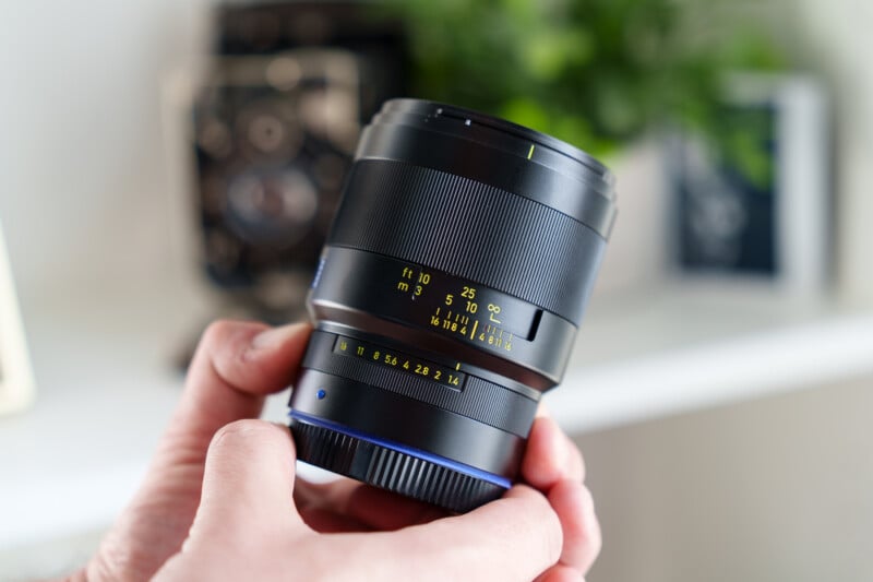 A person holding a camera lens with one hand. The lens features distance markings in feet and meters, with aperture settings visible. The background is blurred, showing a plant and a picture frame.