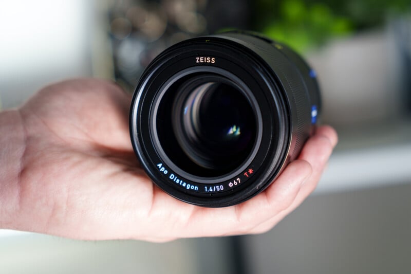A person holds a Zeiss Apo Distagon 1.4/50 camera lens in their hand. The lens features a black body with brand and model details around the glass. A blurred background with green leaves is visible.