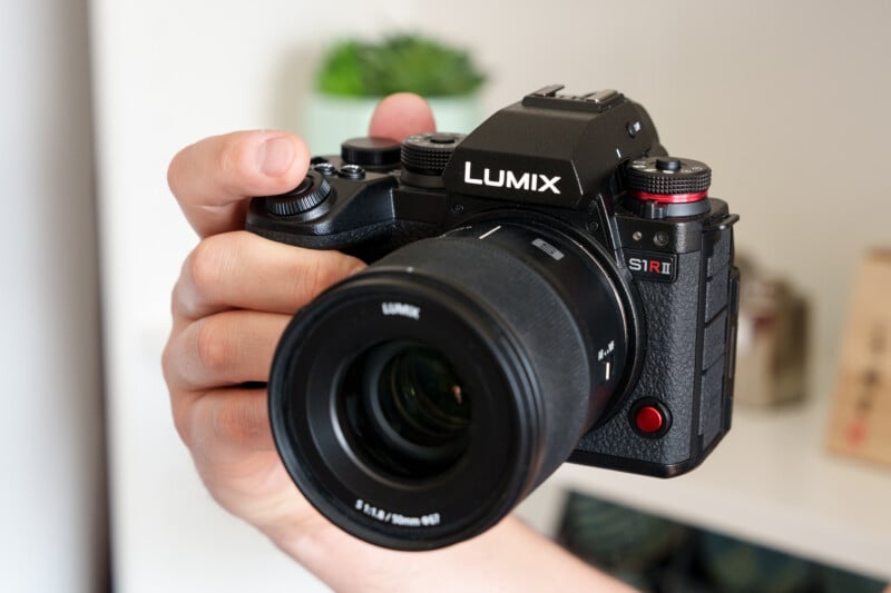 A person holding a black Lumix S1R camera, with fingers gripping the lens. The background is softly blurred, featuring a small plant and shelves.