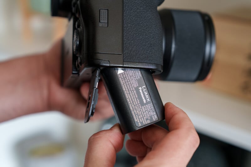 Close-up of hands inserting a Panasonic Lumix battery into a camera. The battery is partially outside of the camera, with visible text and specifications. The background is blurred.