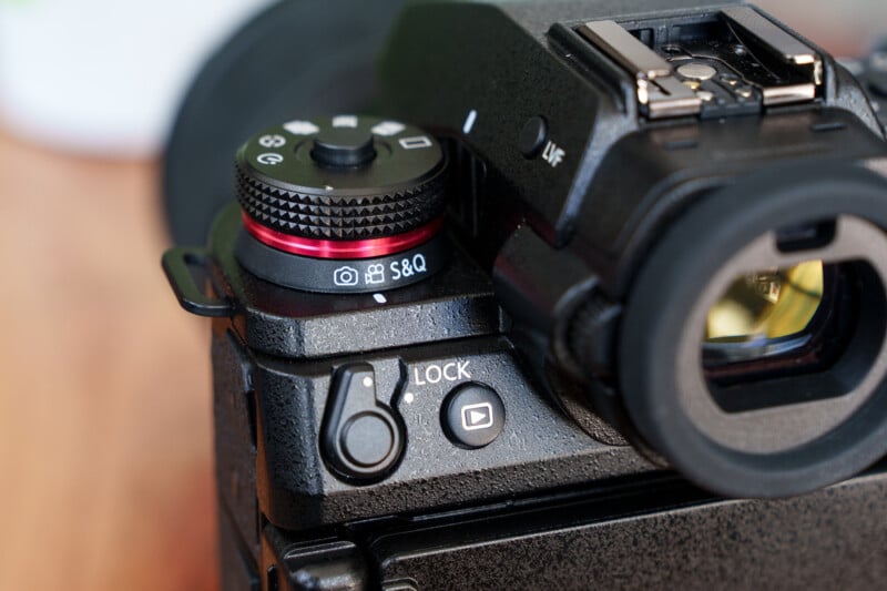 Close-up view of a DSLR camera's control dials and buttons, including the mode dial with settings and a lock switch. The camera body is black with a textured grip and the scene is well-lit.