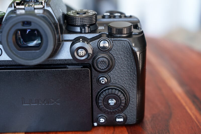 Close-up of the back of a Lumix digital camera on a wooden surface. Visible controls include various buttons, dials, and a partially extended screen, with textured grips and markings.