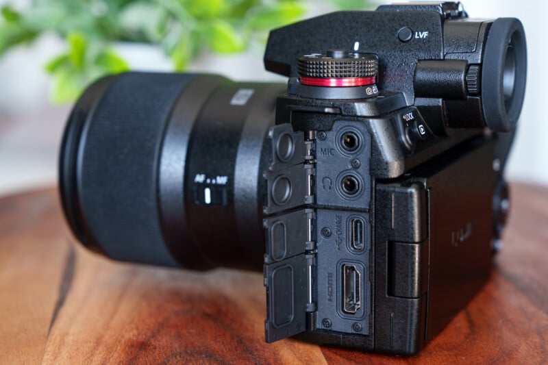 A digital camera with a large lens is positioned on a wooden surface. The side panel is open, revealing various ports including HDMI, microphone, and audio jacks. A plant with green leaves is blurred in the background.