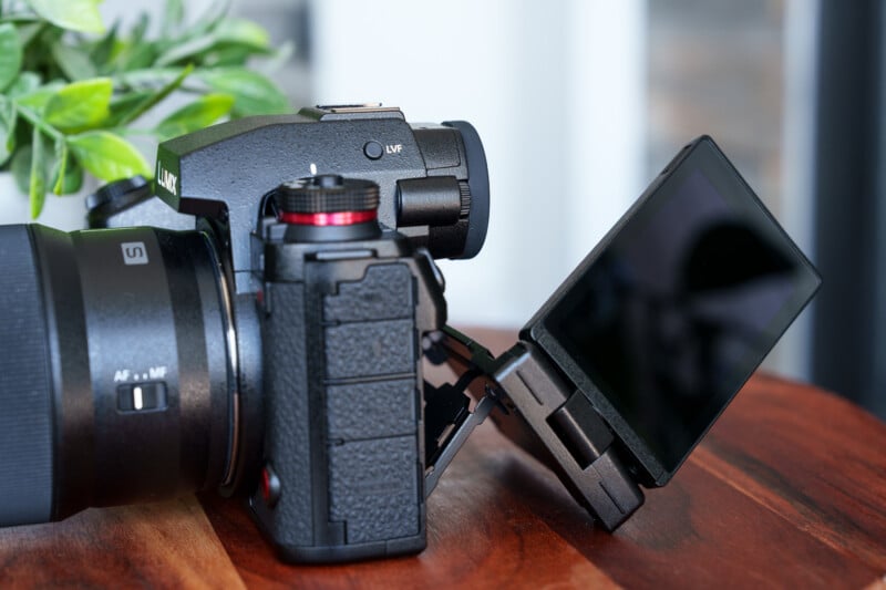 A black digital camera with a fold-out screen is placed on a wooden surface. The camera is partially rotated to show the adjustable LCD screen, lens, and viewfinder. A leafy plant is visible in the background.