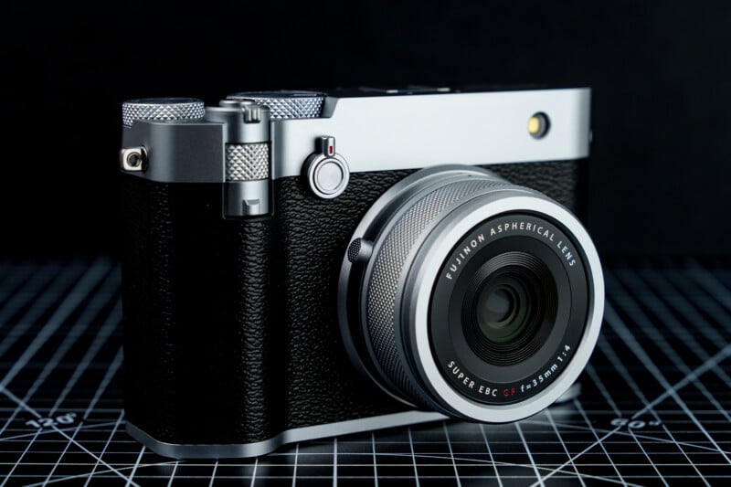 A silver and black camera with a textured grip, featuring a Fujinon aspherical lens, is placed on a black grid surface marked with white lines, set against a dark background.