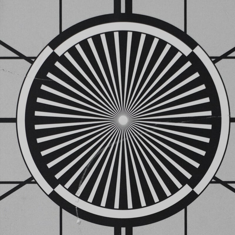 A black and white circular pattern with alternating thick and thin radial lines emanating from the center, resembling a test target, set against a gray background with intersecting straight lines.