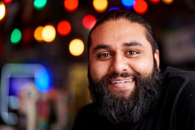 Smiling person with a beard in a black shirt, surrounded by colorful, blurred lights in the background, creating a warm and cheerful atmosphere.