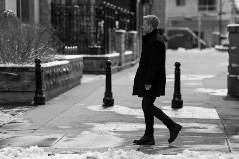 A man in a long coat and boots walks down a snowy, urban street. It's overcast, and patches of snow are scattered on the sidewalk. Bare trees and buildings line the street in the background. The image is in black and white.
