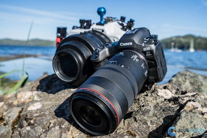 A Canon EOS R5 camera with a large lens rests on a rocky surface by a lakeside. The water and distant shoreline with blurred trees are visible in the background under a clear blue sky.