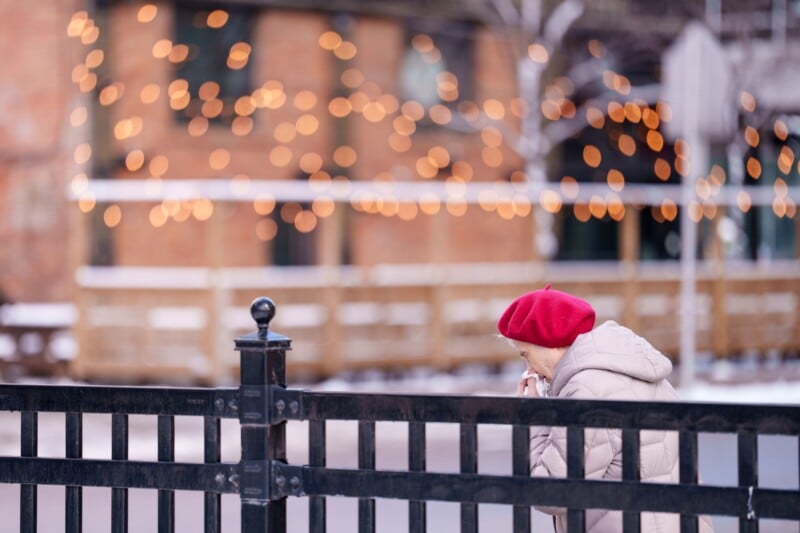 An elderly person wearing a bright red beret and beige coat stands at a black fence, with warm bokeh lights and a brick building softly blurred in the background.