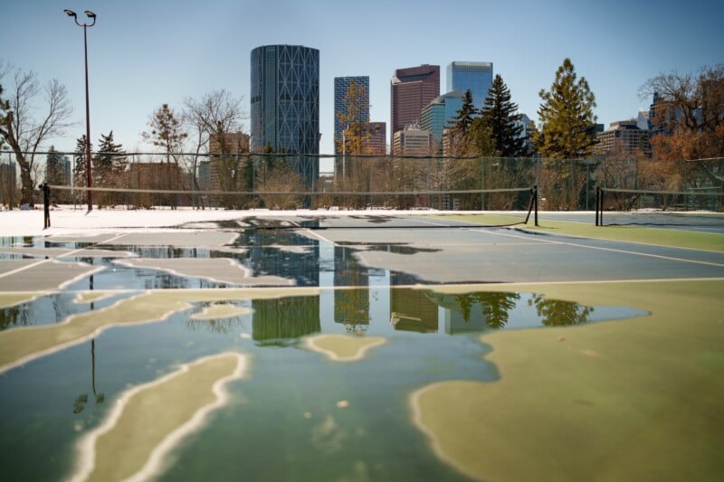 A deserted tennis court with puddles reflecting nearby trees and Calgary's skyline. Snow surrounds the court, contrasting with the clear blue sky. The city's skyscrapers are visible in the background.
