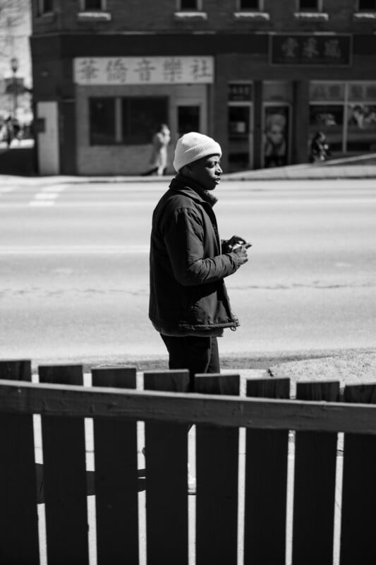 A man wearing a beanie and holding an object stands by a wooden fence on a sidewalk. The background shows a street and a brick building with signs and windows. The image is in black and white.