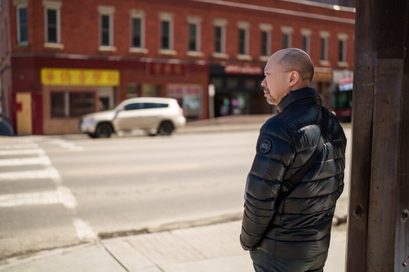 A man in a black puffer jacket stands on a sidewalk looking across an empty street. In the background, there's a red brick building with Asian signage and a white SUV parked on the street.