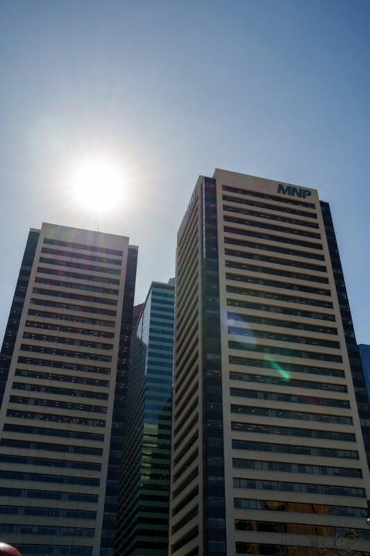 Two tall office buildings with numerous windows stand against a blue sky. The sun is shining brightly near the top left, creating lens flare effects. The building on the right has "MNP" written near the top.