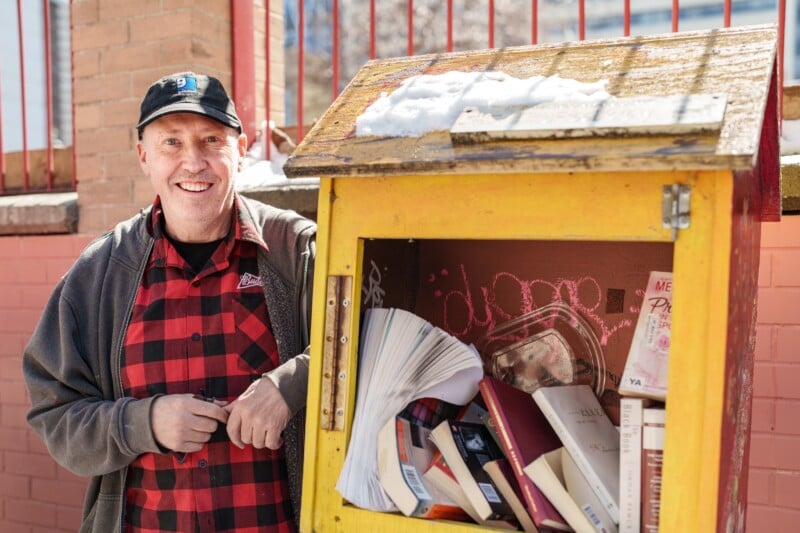 Smiling man in a black cap and red plaid shirt stands beside a bright yellow free library box filled with books and papers. Snow is on top of the box, and a red brick wall is in the background.