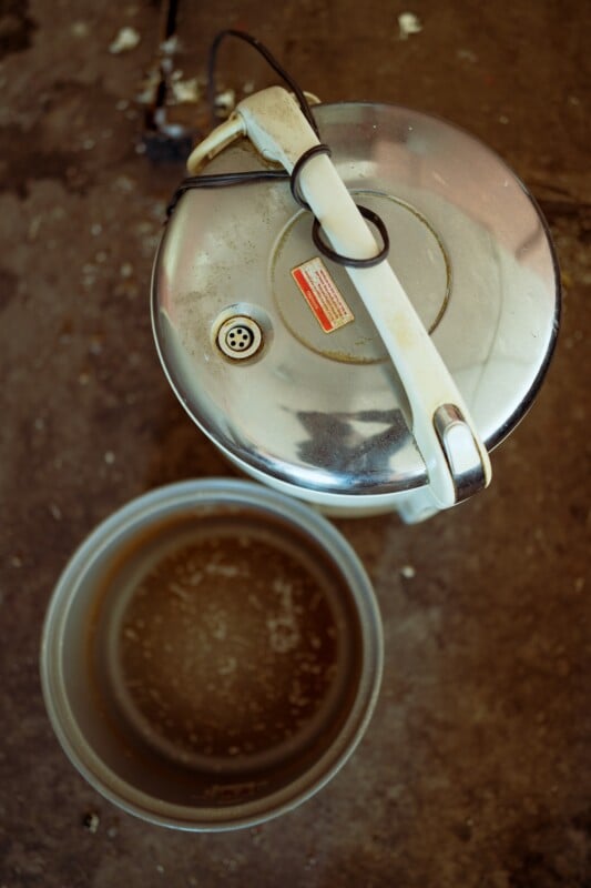 Top view of a stainless steel rice cooker with a lid and a detached inner pot on a worn, dark surface. The handle is secured with a cord, and the inner pot shows signs of use, with stains visible on its bottom.