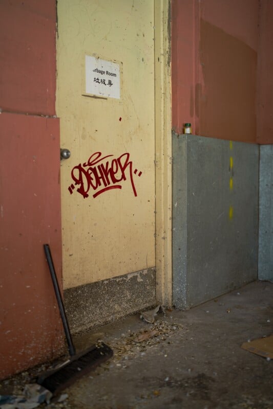 A weathered door marked "Garbage Room" with a red graffiti tag is set in a wall of peeling paint. A broom leans against the door, and debris litters the ground. A small bottle is on a ledge to the right.