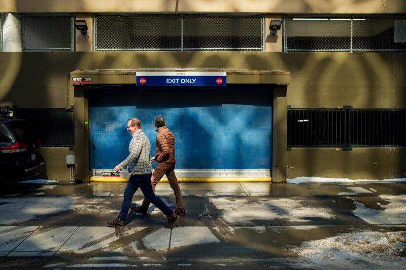 Two people walk past a snow-covered sidewalk in front of a blue "Exit Only" garage door. They wear winter jackets, reflecting the bright sunlight casting shadows from above. A car is partially visible on the left.