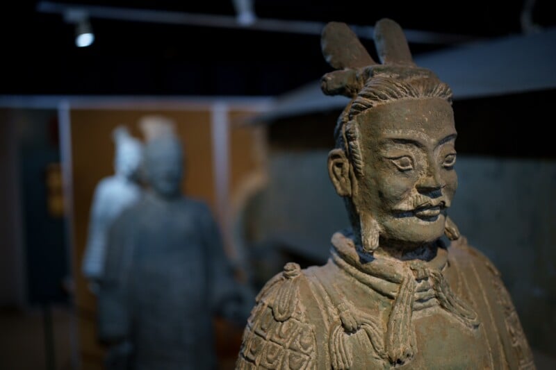 Close-up of a terracotta warrior statue, showing detailed facial features and armor. In the background, other figures are blurred. The image captures the intricacy and artistry of these ancient sculptures.