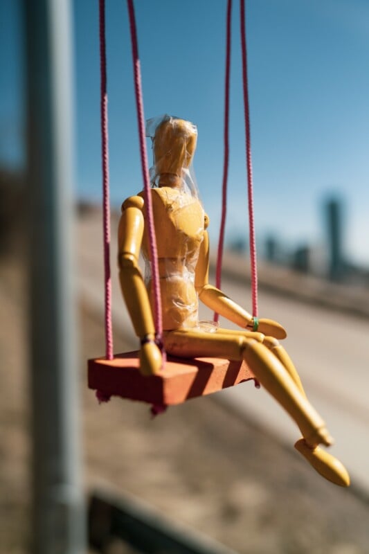 A wooden mannequin wrapped in plastic sits on a small red swing with pink ropes. The swing is set against a blurred outdoor background with a clear blue sky and indistinct buildings in the distance.