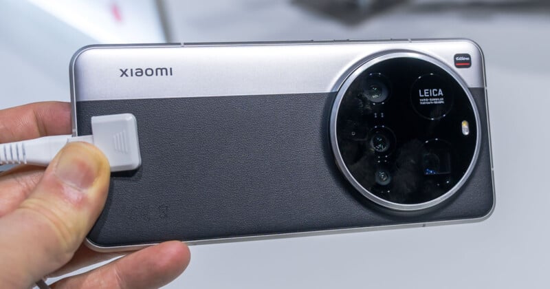 A person holds a Xiaomi smartphone showcasing its back side. The phone features a large circular camera module branded with "Leica," containing multiple lenses. The design is sleek with a black and silver finish.