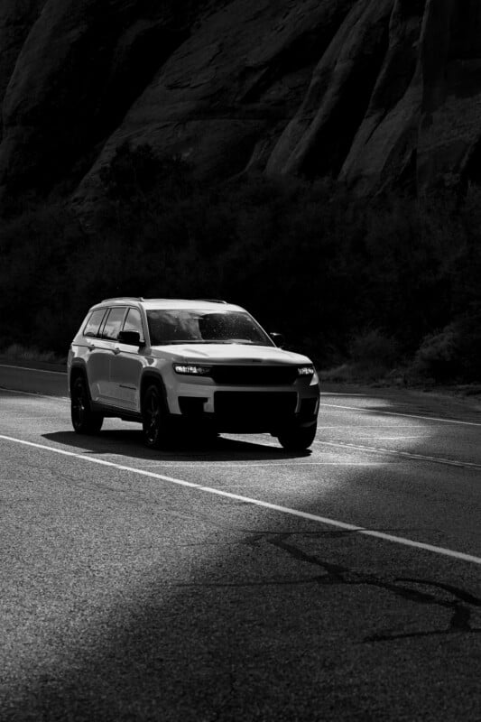 A black and white image of an SUV driving on a winding road. Sunlight highlights the vehicle as it passes through a shadowed area, with rocky cliffs visible in the background.