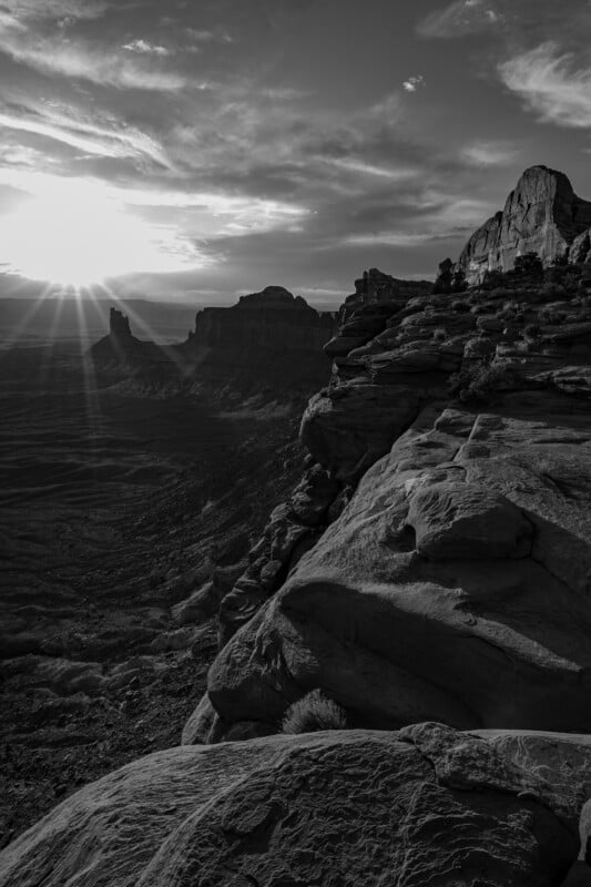 Black and white image of a rocky cliff overlooking a vast landscape with distant mesas. The sun is setting on the horizon, casting sunrays and dramatic shadows across the scene.