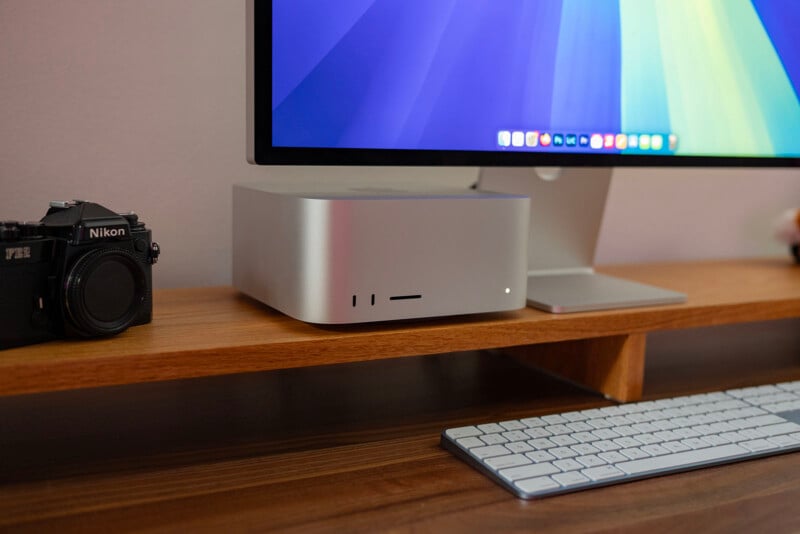 A desktop setup featuring a large monitor with a colorful gradient screen, a compact silver computer, and a keyboard. A vintage Nikon camera sits to the left on a wooden shelf. The setting is minimalistic and organized.
