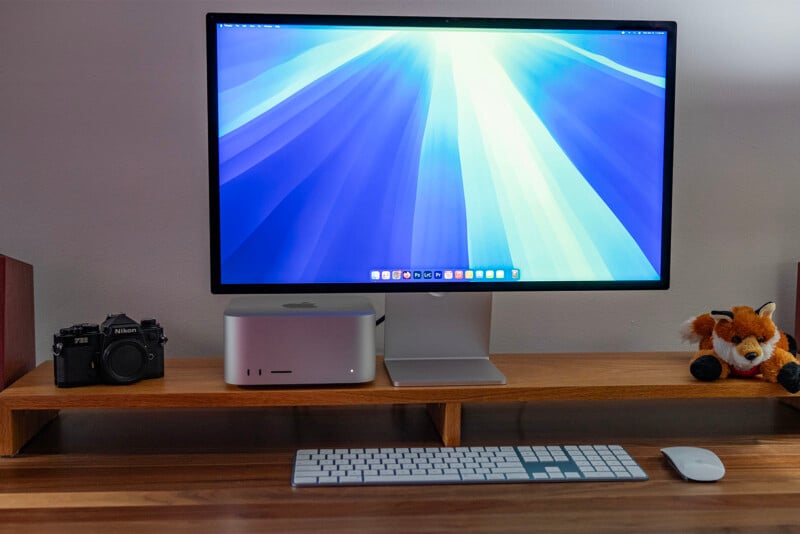 A tidy desk setup featuring a large monitor displaying a blue and white abstract background. Below, there's a keyboard and mouse. To the left, a camera, and to the right, a plush fox toy rests on a wooden shelf.