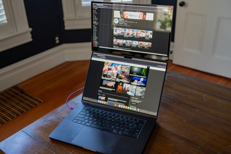 A laptop on a wooden table displays a YouTube channel, while an external monitor above shows a similar YouTube interface. The setup is in a room with dark walls, a window, and a white door in the background.