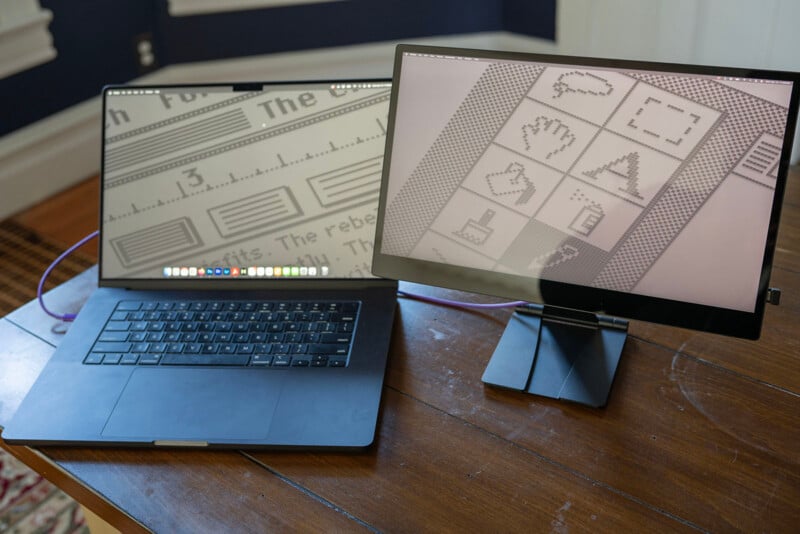A sleek laptop and a separate monitor display retro pixelated graphics resembling an old operating system. The laptop shows a text editor, while the monitor displays icons. Both devices are on a wooden table.