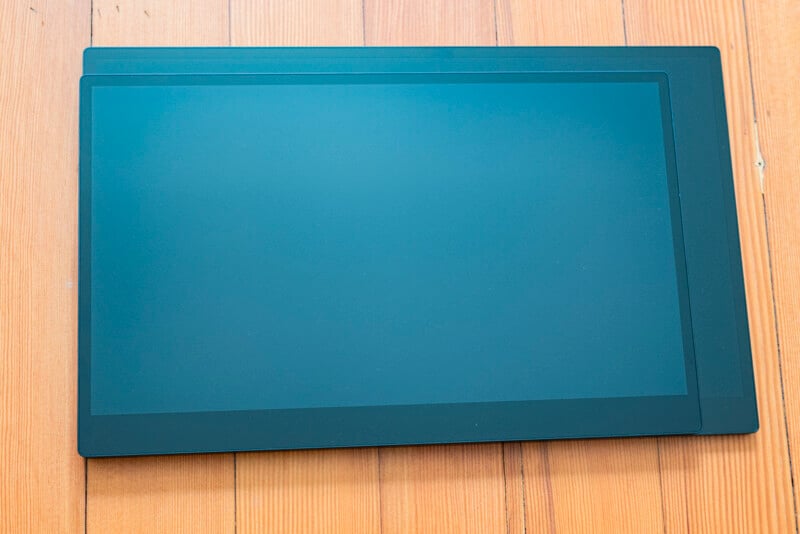 A large, flat rectangular electronic device, possibly a tablet or monitor, lies on a wooden surface. The screen is blank and the device has a thin black border.