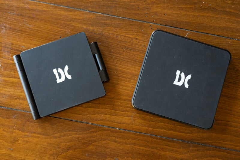 Two compact, black digital cameras with a foldable design are displayed on a wooden surface. Both cameras feature the same white logo on their covers and vary slightly in shape, one being more rectangular and the other square.
