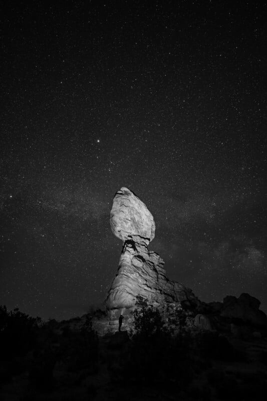 A black and white image of a large, balanced rock formation under a starry night sky. A person stands at the base, silhouetted against the illuminated rock, surrounded by bushes and rocky terrain.