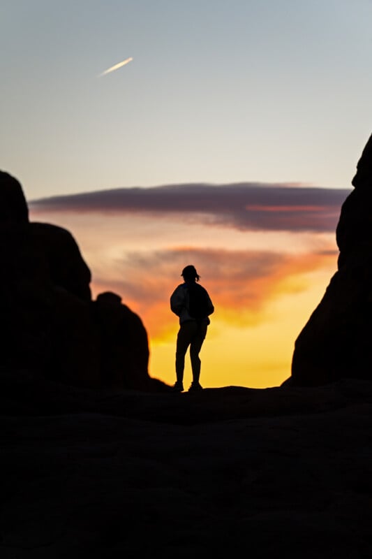 Silhouette of a person standing between rock formations against a vibrant sunset sky. The sky features shades of orange, pink, and purple with a small streak of a contrail visible above.