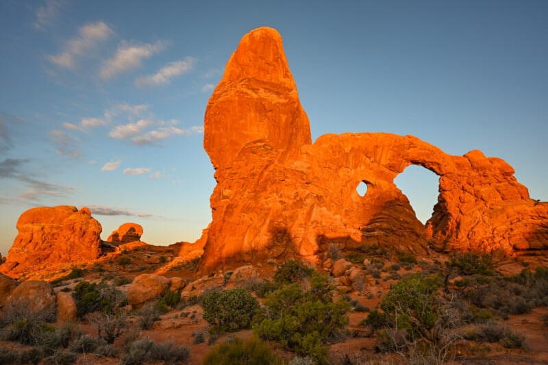 Rock formations at Arches National Park bask in warm, golden sunlight during sunset. The rough terrain is dotted with shrubs, and the sky is clear with a few scattered clouds, highlighting the natural arch and towering structures.