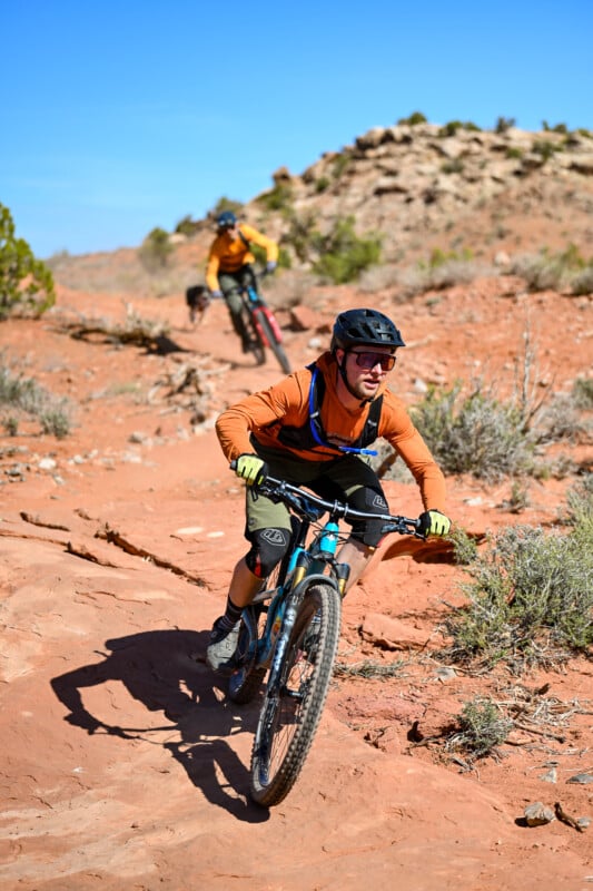 Two people mountain biking on a rocky desert trail under a clear blue sky. The leading cyclist wears an orange shirt and helmet, while the second rider follows closely behind. Rugged terrain and distant hills are visible in the background.