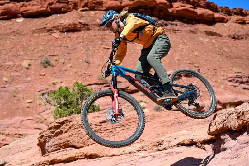 A person wearing a helmet and backpack rides a mountain bike off a rocky ledge in a desert landscape, set against red rock formations and a clear blue sky.