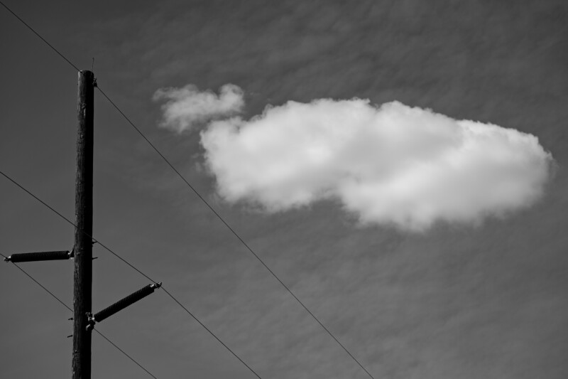 Black and white image of a single fluffy cloud beside a wooden utility pole with power lines extending across the sky.