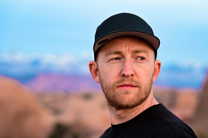 A man with a beard and a black cap gazes to the side against a blurred backdrop of rocky terrain and distant snow-capped mountains. The sky is clear and blue, giving a serene atmosphere.