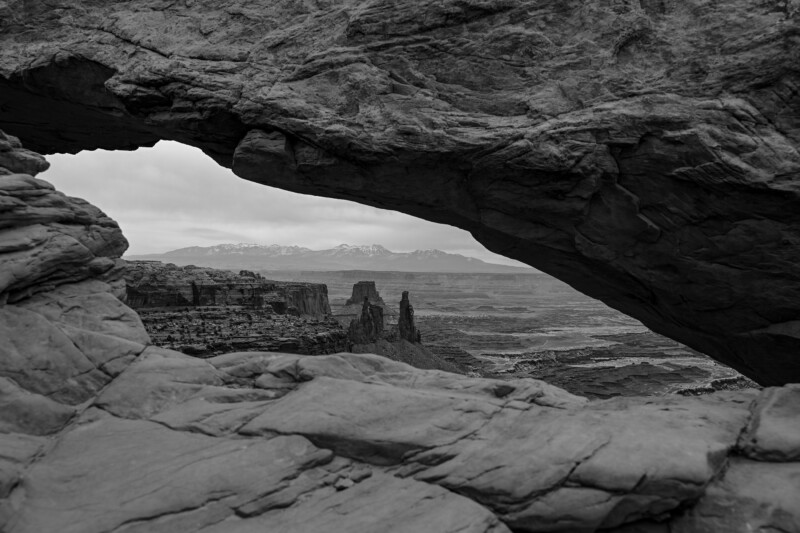 A black and white photo of a natural rock arch overlooking a desert landscape with mesas and distant snow-capped mountains under a cloudy sky. The arch frames the rugged, expansive scenery.